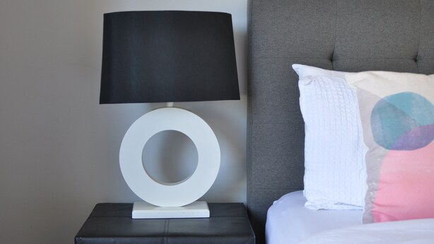 A modern bedroom with a grey upholstered headboard, a white circular bedside table, a black lamp, and a white pillow with a colorful design. - Modern Home near Swan Valley (Beechboro)