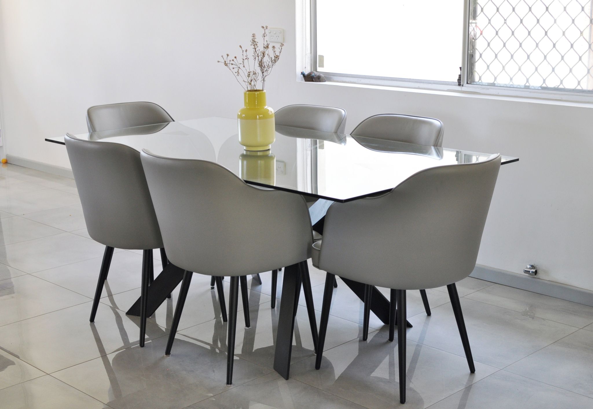 A modern dining area with a glass table surrounded by four grey chairs with black legs.