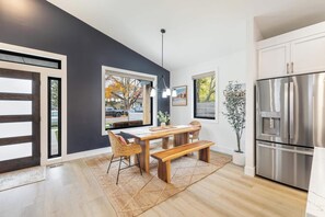 Eine moderne Küche mit einem großen Edelstahl-Kühlschrank, einem Essbereich mit einem Holztisch und Stühlen sowie Blick ins Freie durch ein Fenster.