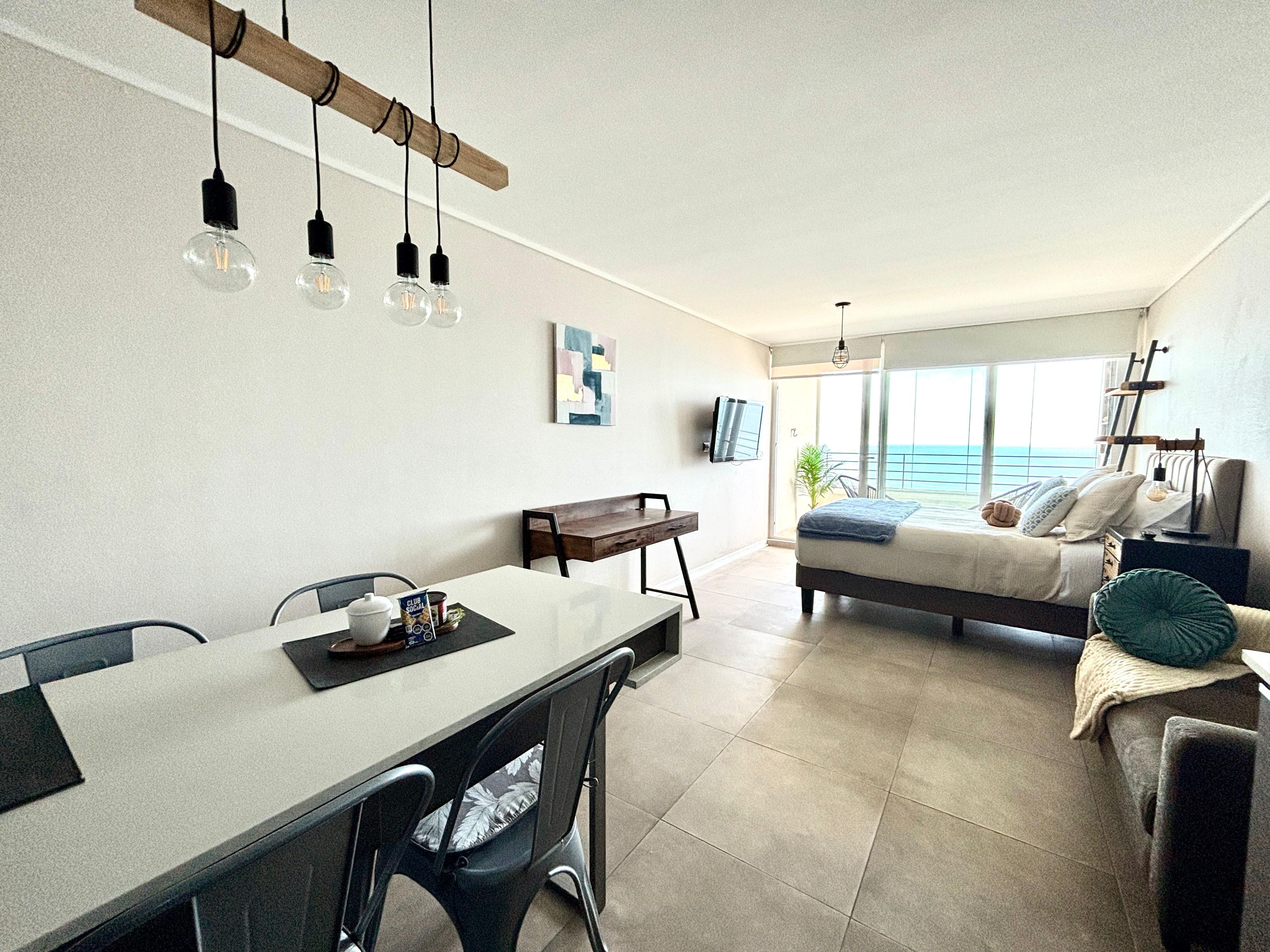 A modern living room with a large bed, a dining area with a table and chairs, and a balcony with a view of the ocean.