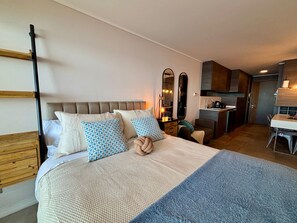 A modern bedroom with a large bed, a bedside table, and a small kitchenette in the background. - Cozy 1 bedroom apartment with a sea view + heated pool + parking in Reñaca. (Viña del Mar)