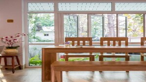 A dining area with a wooden table and benches, large windows with blinds, and a small side table with a vase of flowers. - Baan I Inspire (Mueang Phrae District)