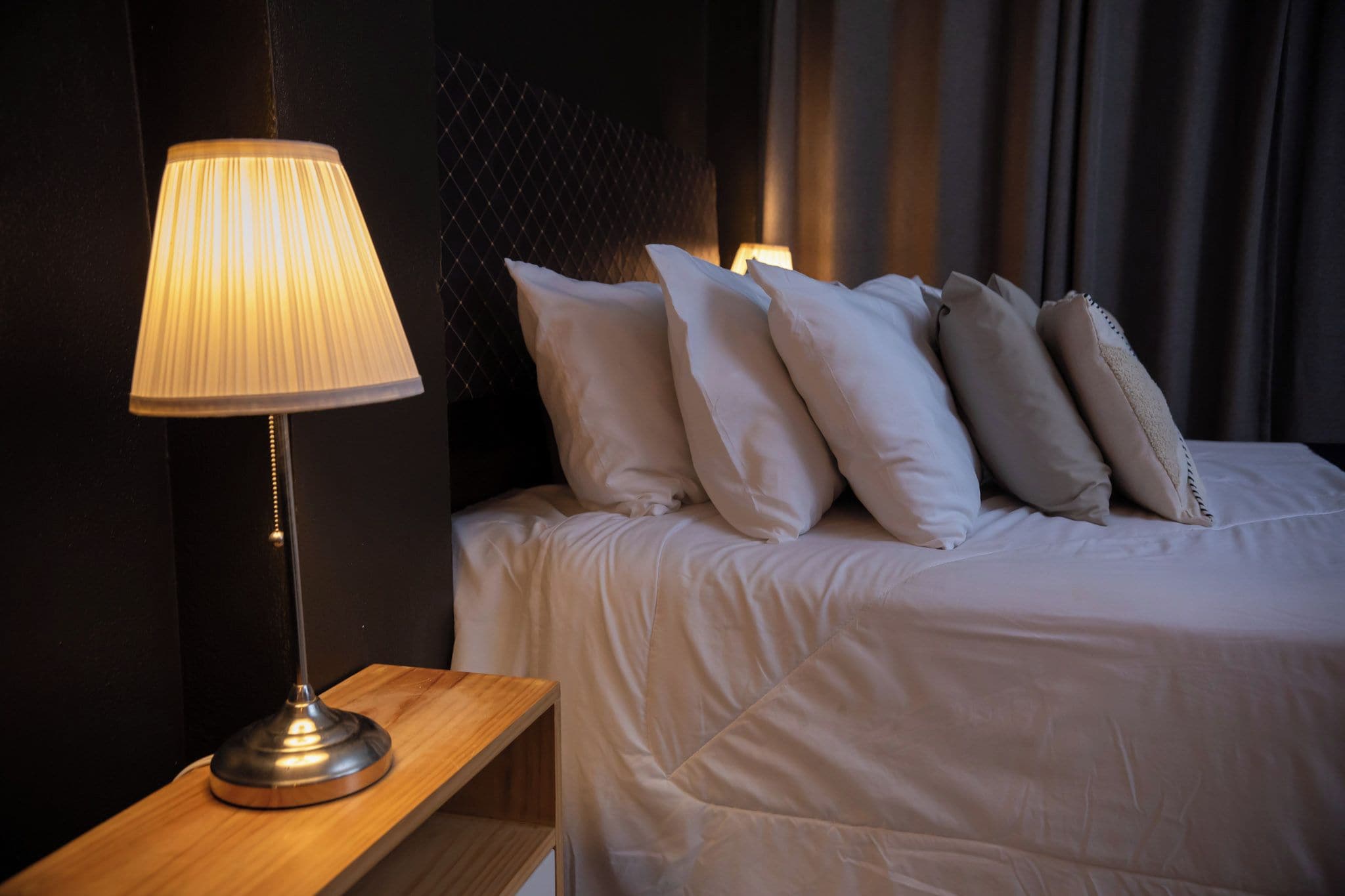 A neatly made bed with white bedding and several pillows, a bedside table with a lamp, and a dark wall with a textured pattern.