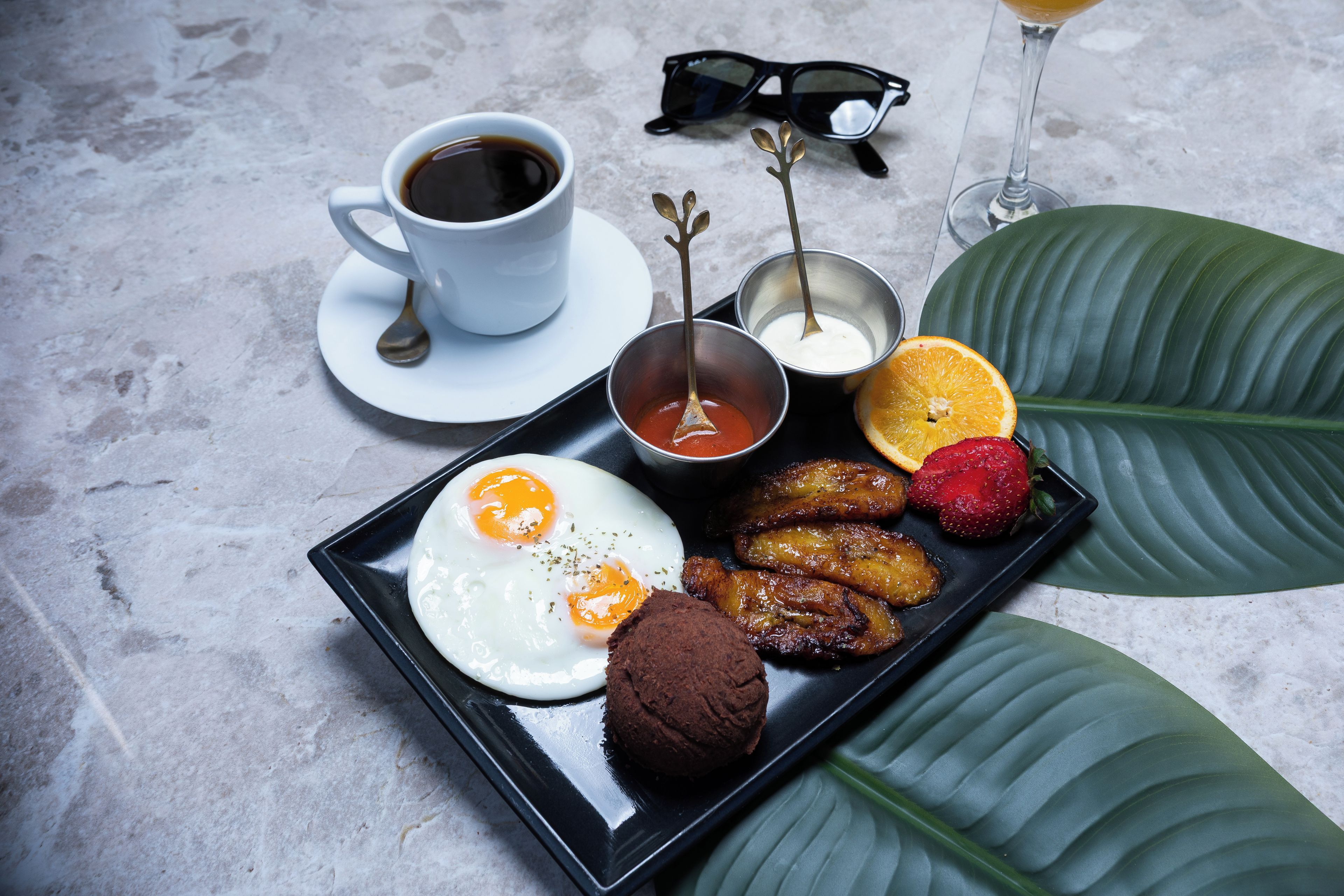 A tray with eggs, sausages, a brownie, and fruit on a table with a cup of coffee and a glass of drink.