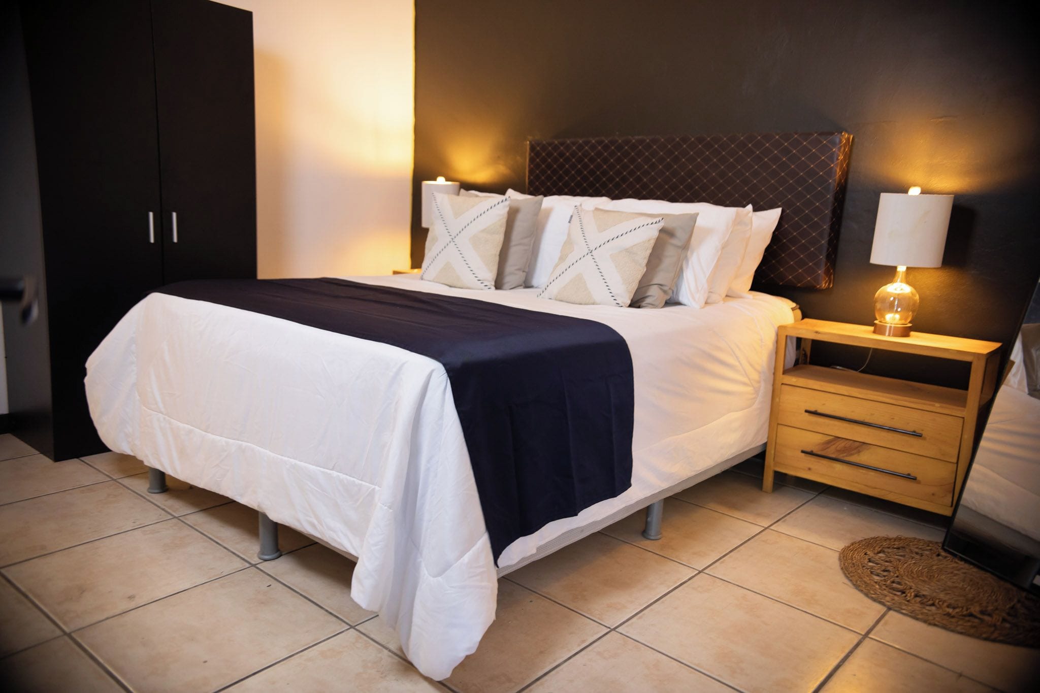 Una cama bien hecha con una colcha azul oscuro y ropa de cama blanca, una mesita de noche de madera con una lámpara y un armario oscuro.