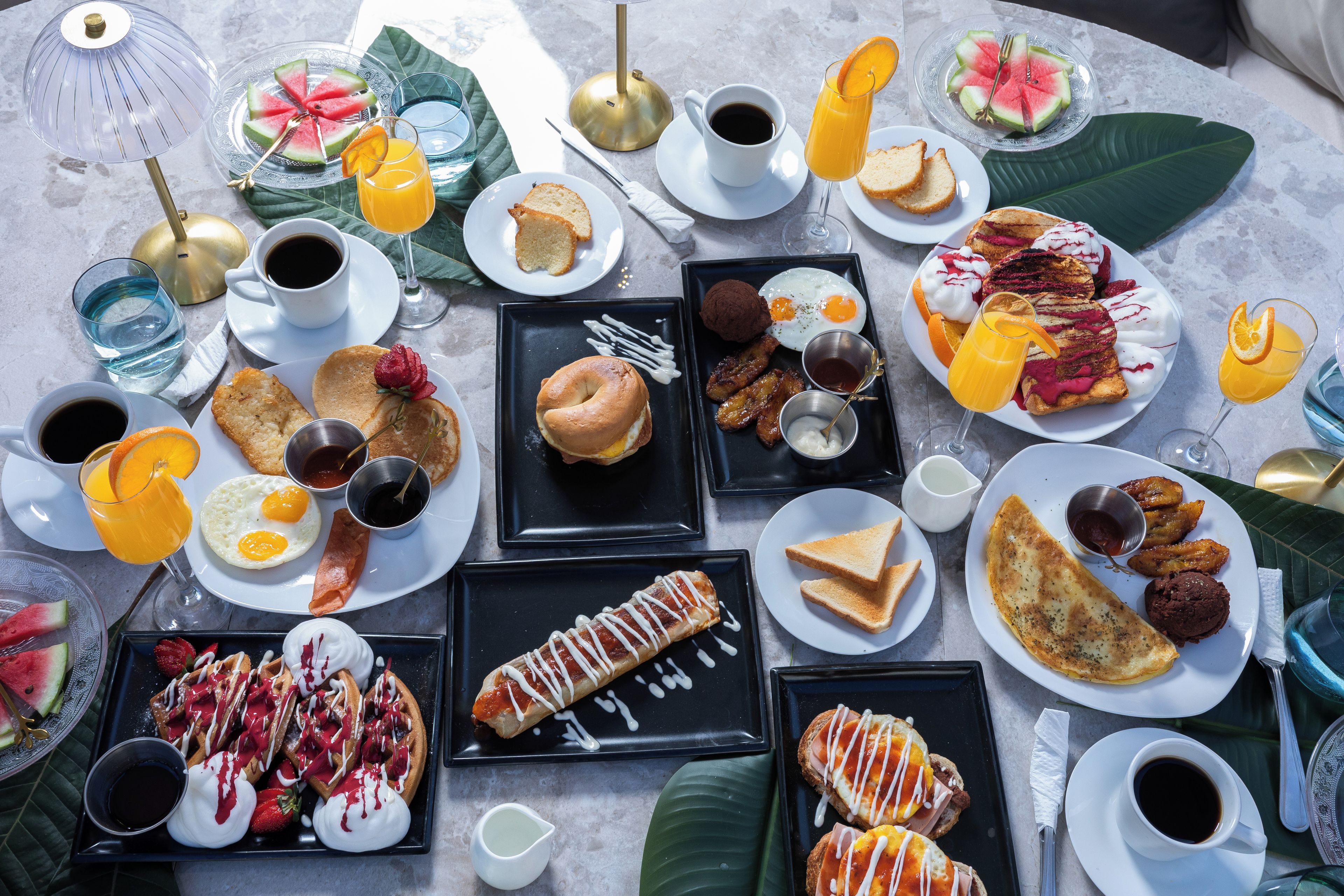 A variety of breakfast items including pancakes, eggs, waffles, and pastries, arranged on multiple plates and trays.
