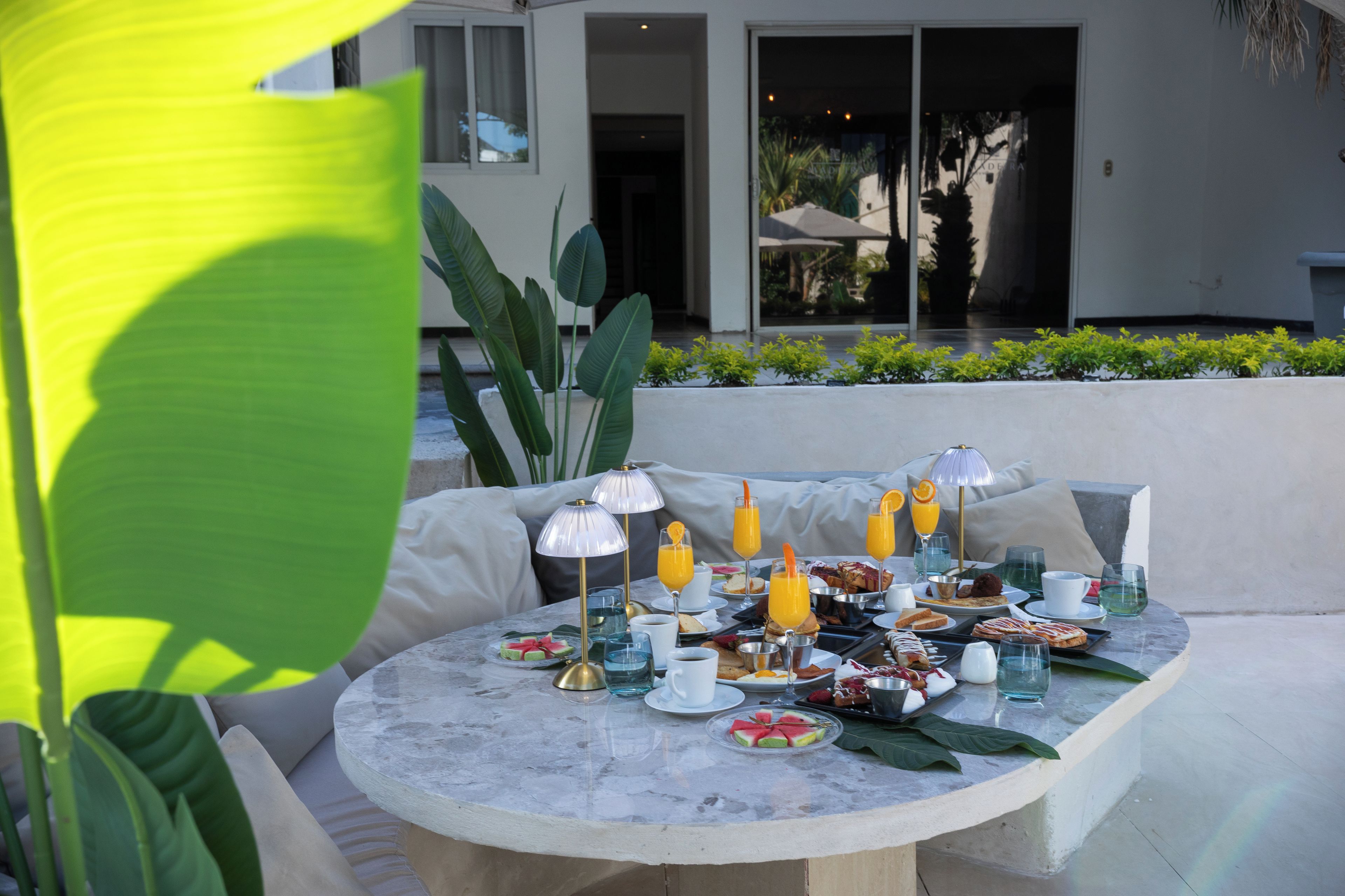 An outdoor dining area with a round table set for breakfast, surrounded by a modern building and greenery.