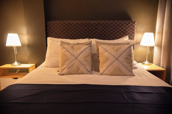 A neatly made bed with two pillows, a dark bedspread, and two bedside lamps on wooden nightstands.