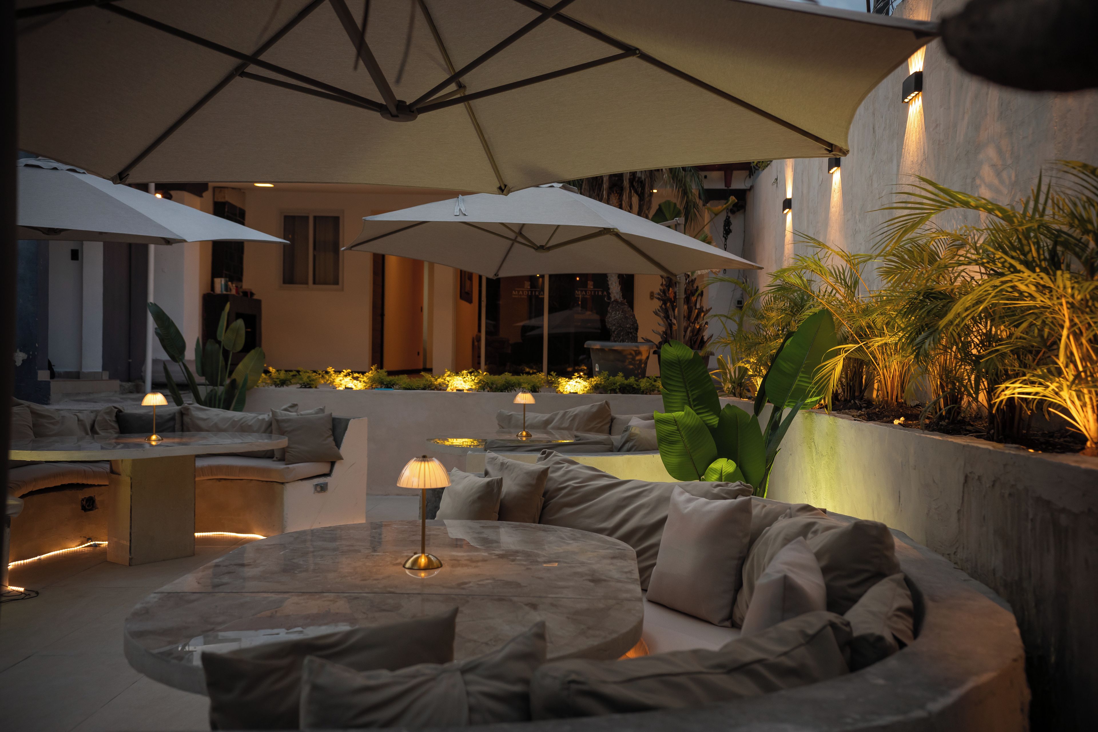 An outdoor seating area with round tables and cushioned benches, shaded by large umbrellas.