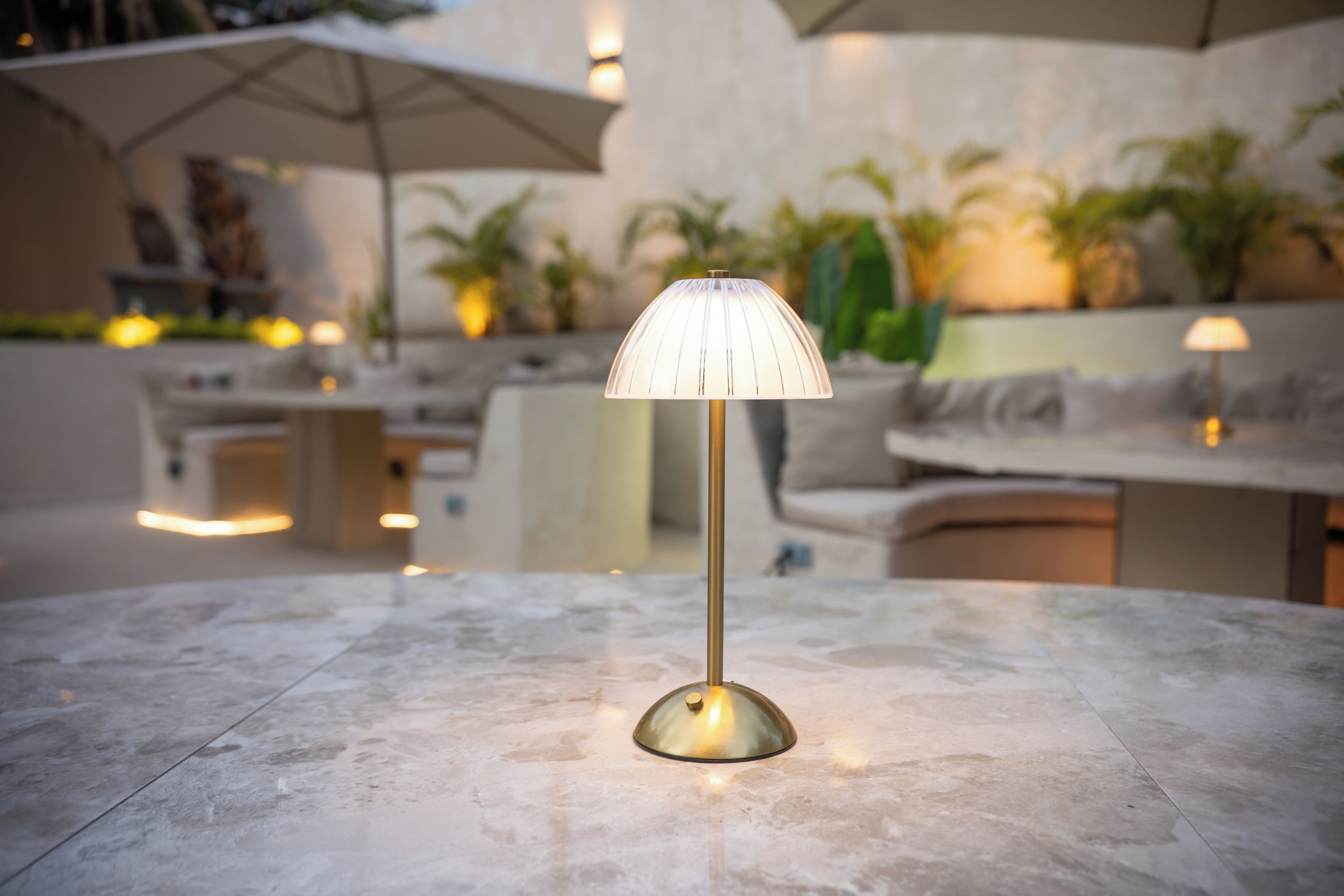 Una mesa con una lámpara moderna en un área exterior bien iluminada, con sombrillas y plantas.