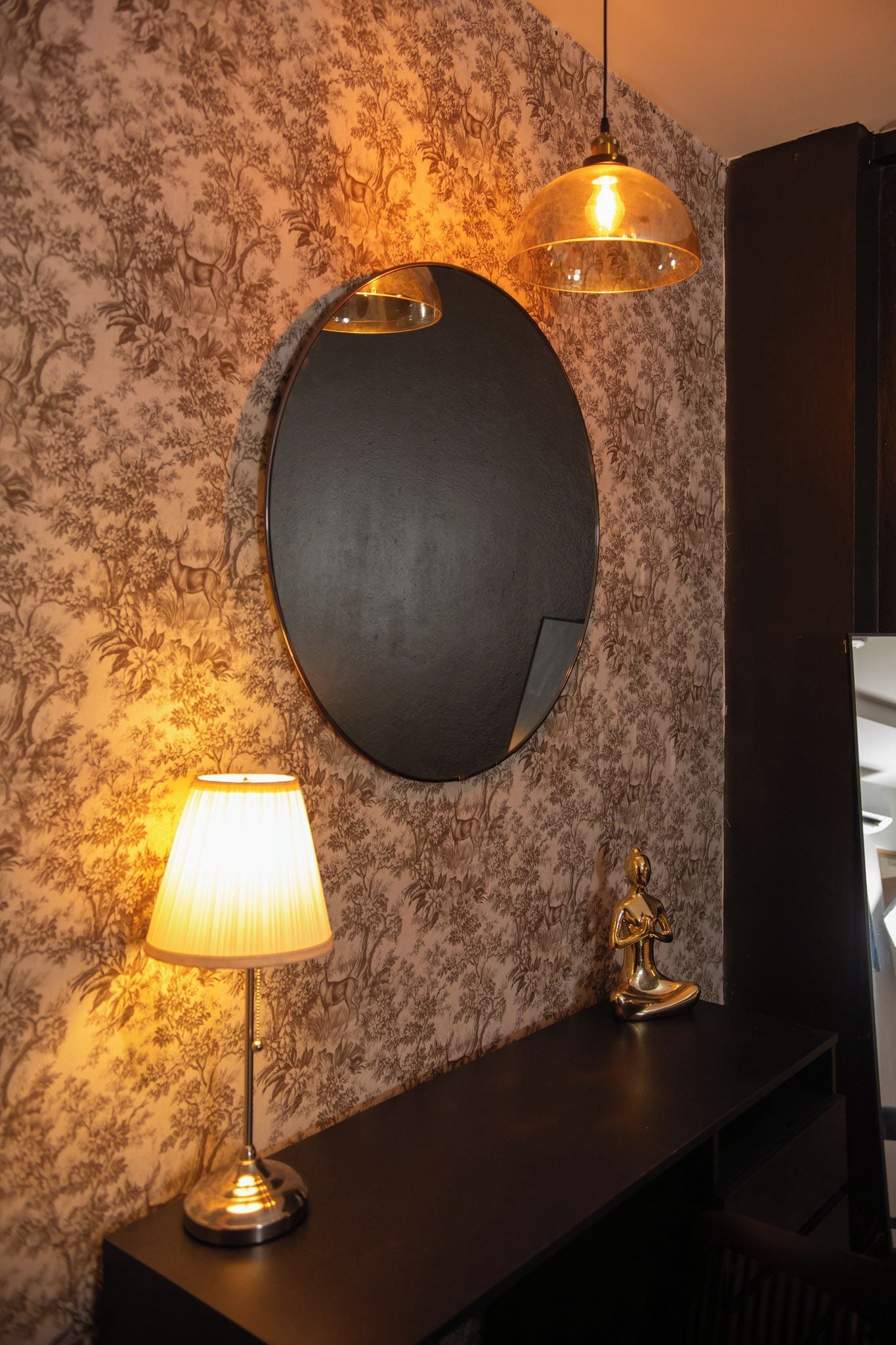 A room with a round mirror, a lamp, and a statue on a dark console.