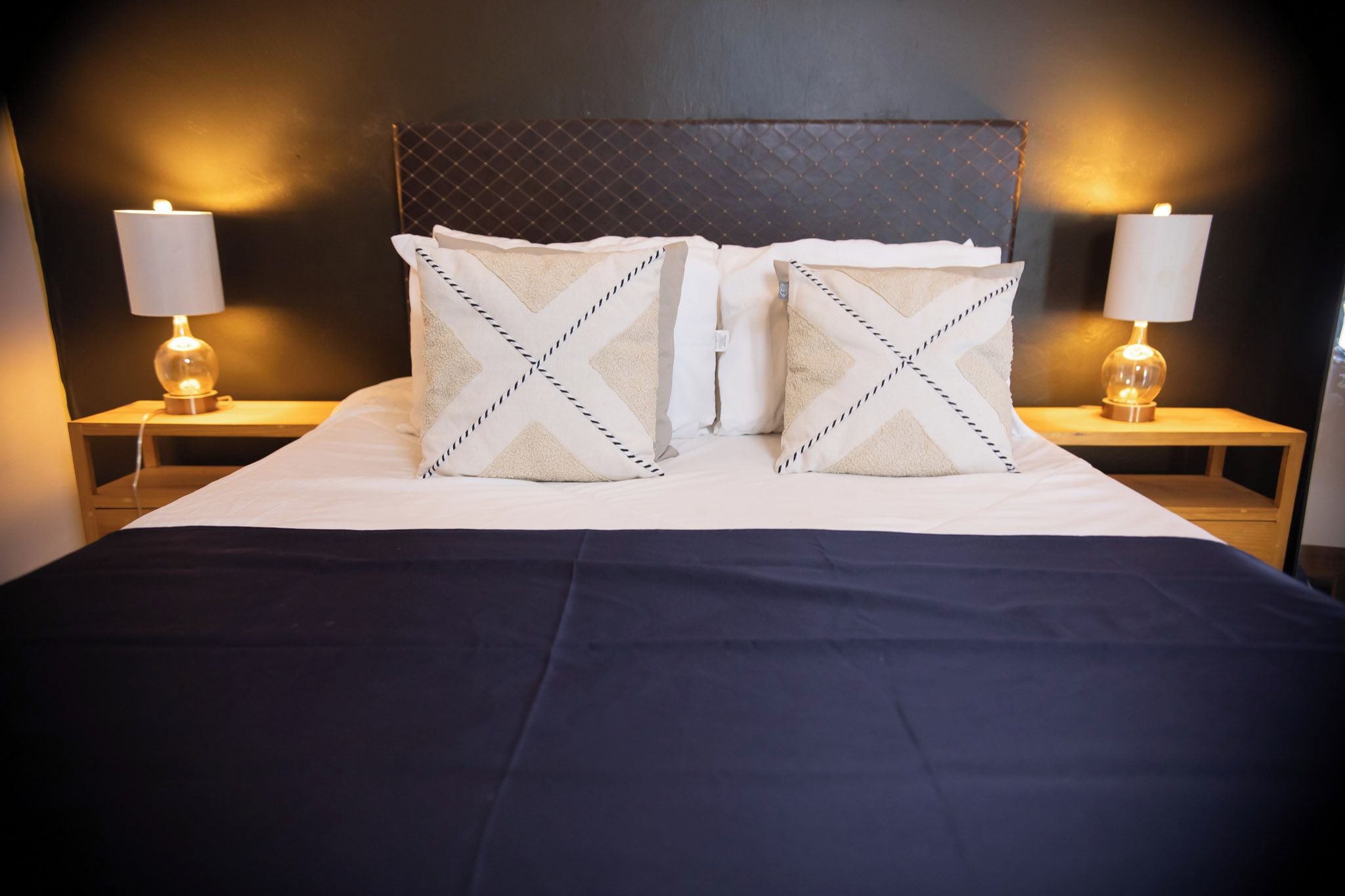 Un lit bien fait, recouvert d’une couvre-lit sombre, avec deux oreillers à motifs chevrons et deux lampes de chevet posées sur des tables de nuit en bois.