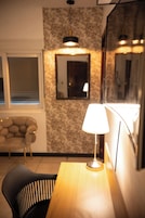 Um quarto com uma mesa de madeira, uma lâmpada, uma cadeira, um sofá e uma parede com um espelho e uma janela.