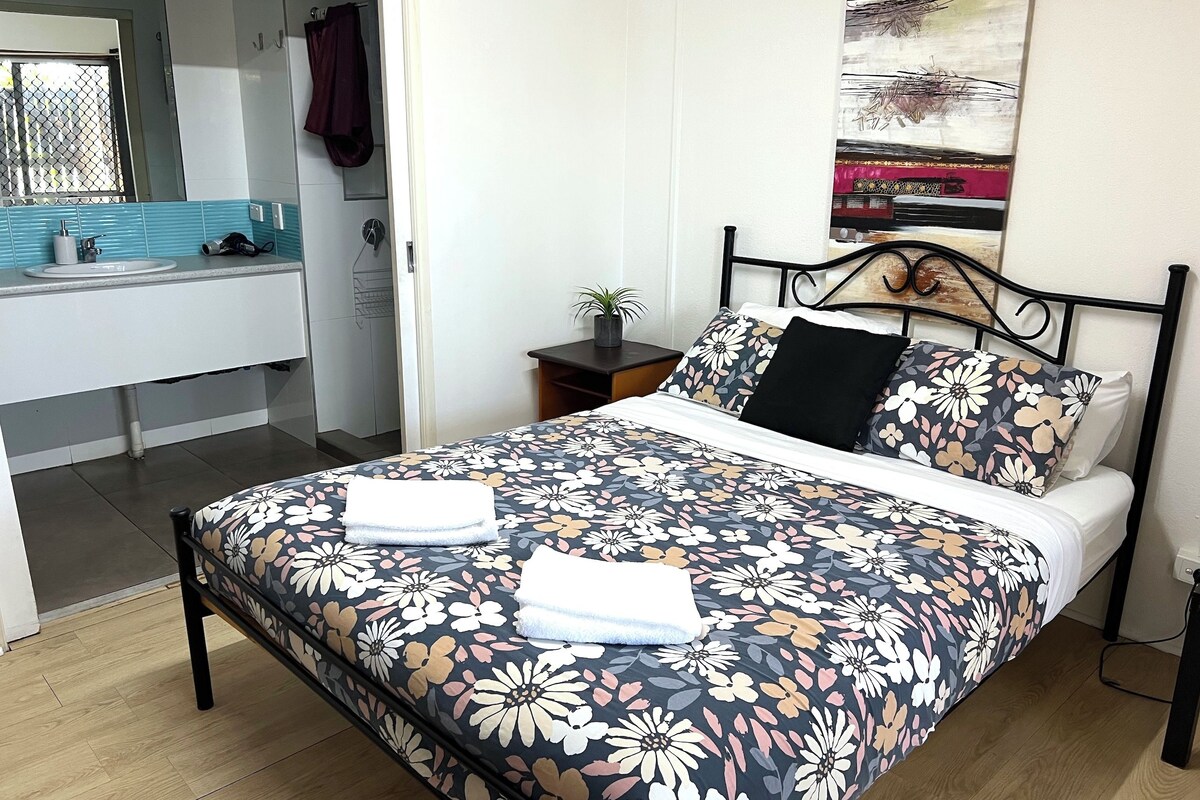 A bedroom with a bed, a nightstand, a potted plant, and a bathroom visible in the background.