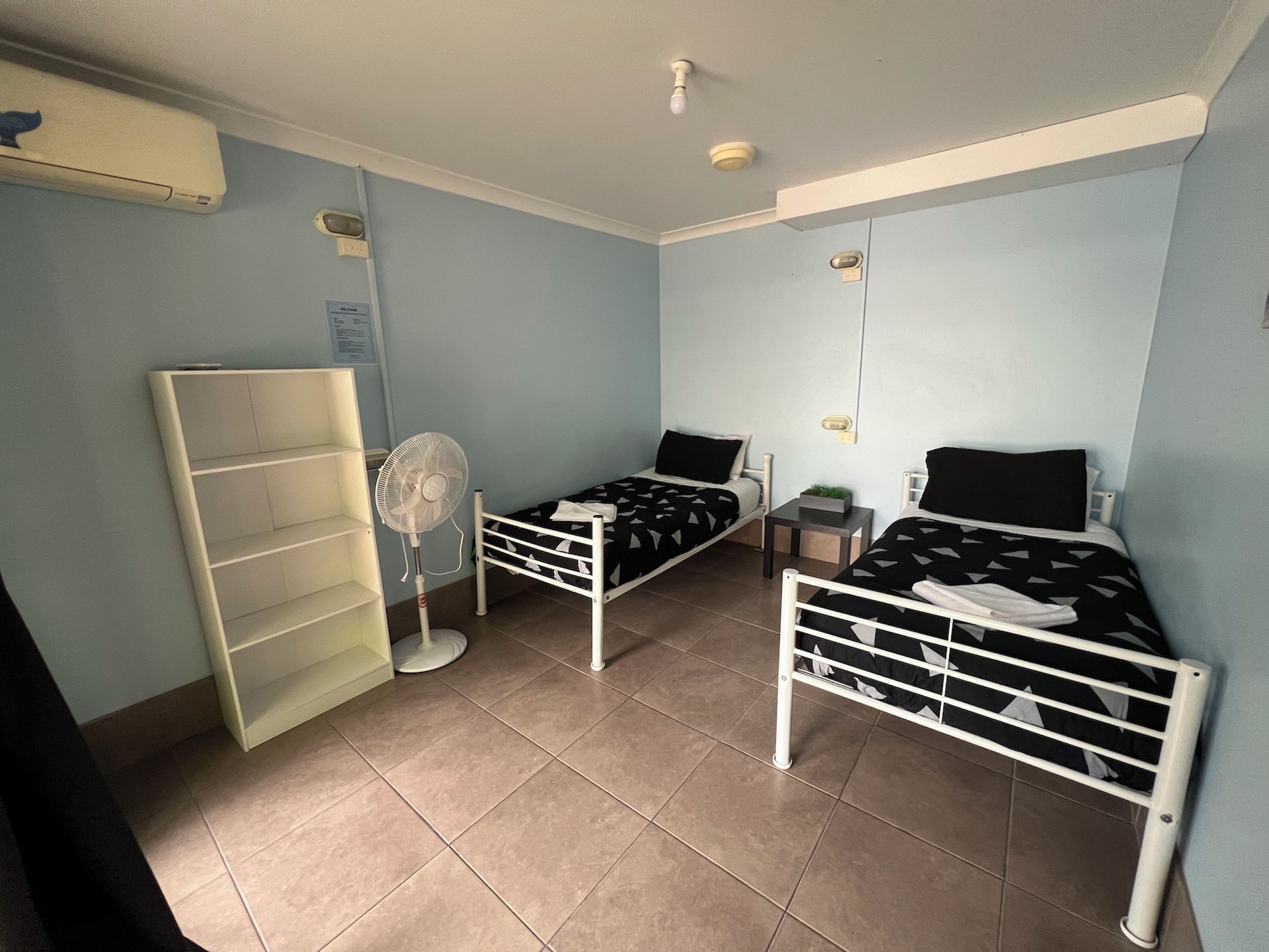 Een kleine, goed verlichte kamer met twee eenpersoonsbedden, een ventilator, een plankenunit en een klein tafeltje.