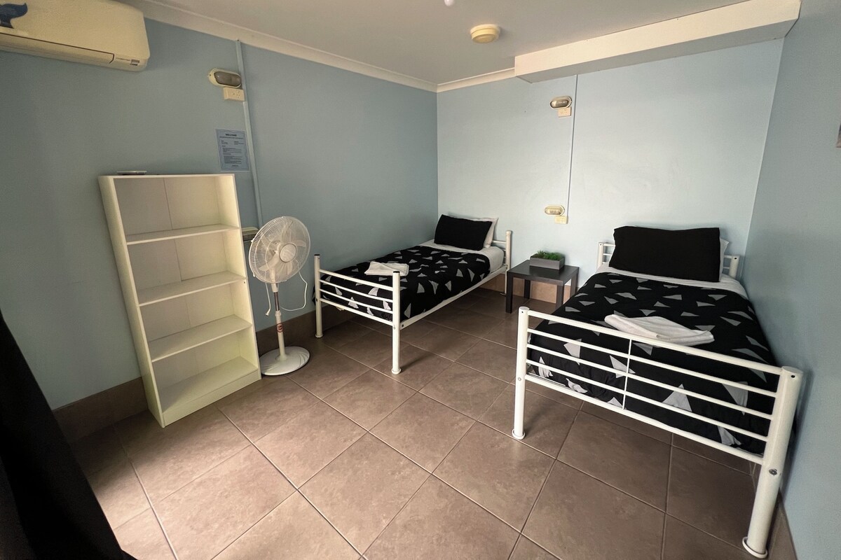 A small, well-lit room with two single beds, a standing fan, a shelf unit, and a small table.