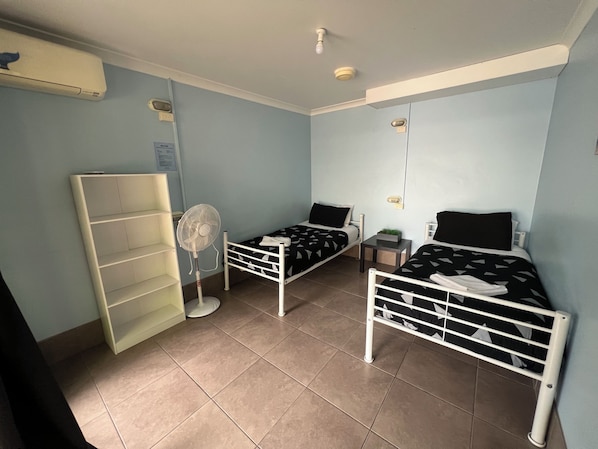 A small, well-lit room with two single beds, a standing fan, a shelf unit, and a small table. - Flashpackers Hervey Bay (Torquay)