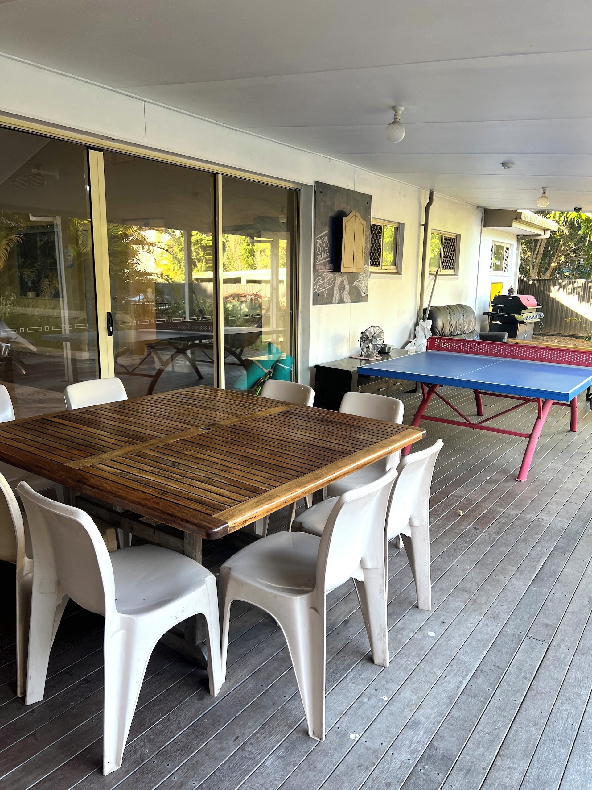 Un patio exterior con mesa de madera y sillas de plástico blanco, mesa de ping pong y una parrilla.