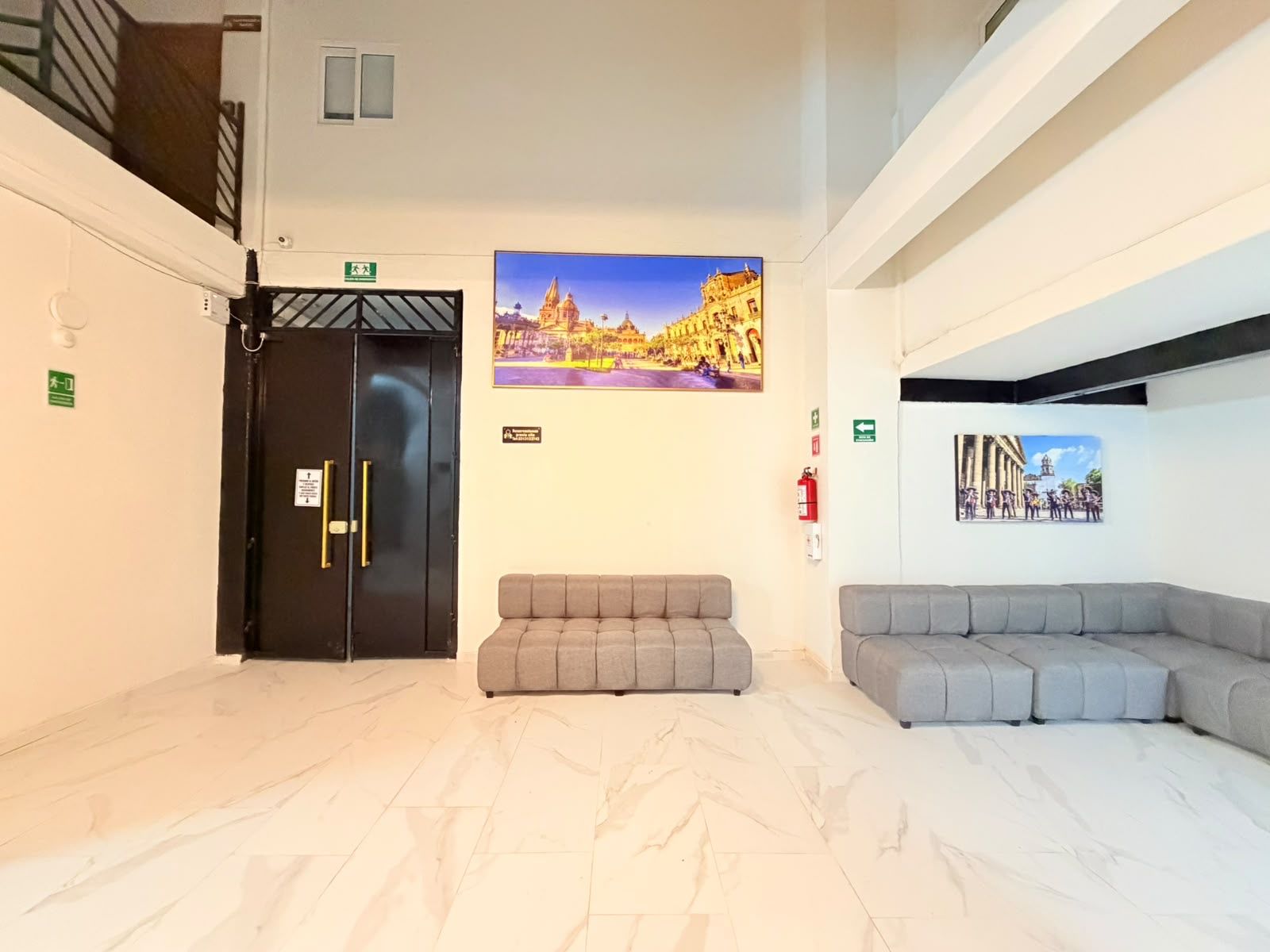 A hotel lobby with a reception desk, a black elevator, grey seating area, and two wall-mounted pictures of buildings.
