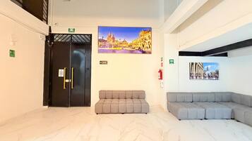A hotel lobby with a reception desk, a black elevator, grey seating area, and two wall-mounted pictures of buildings.