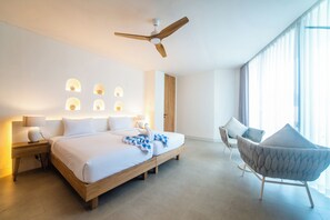A modern hotel room with a large bed, a ceiling fan, a seating area with a chair, and a large window with curtains. - Kona Resort Pererenan F (Badung)