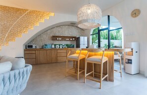 A modern kitchen with a stone backsplash, wooden bar stools, and a large window providing a view of greenery. - Kona Resort Pererenan F (Badung)