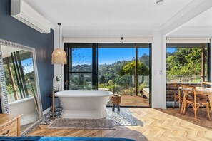 A modern bathroom with a freestanding bathtub, a large mirror, and a view of a lush landscape through glass doors. - Fletchers Ridge Guest House (Montville)