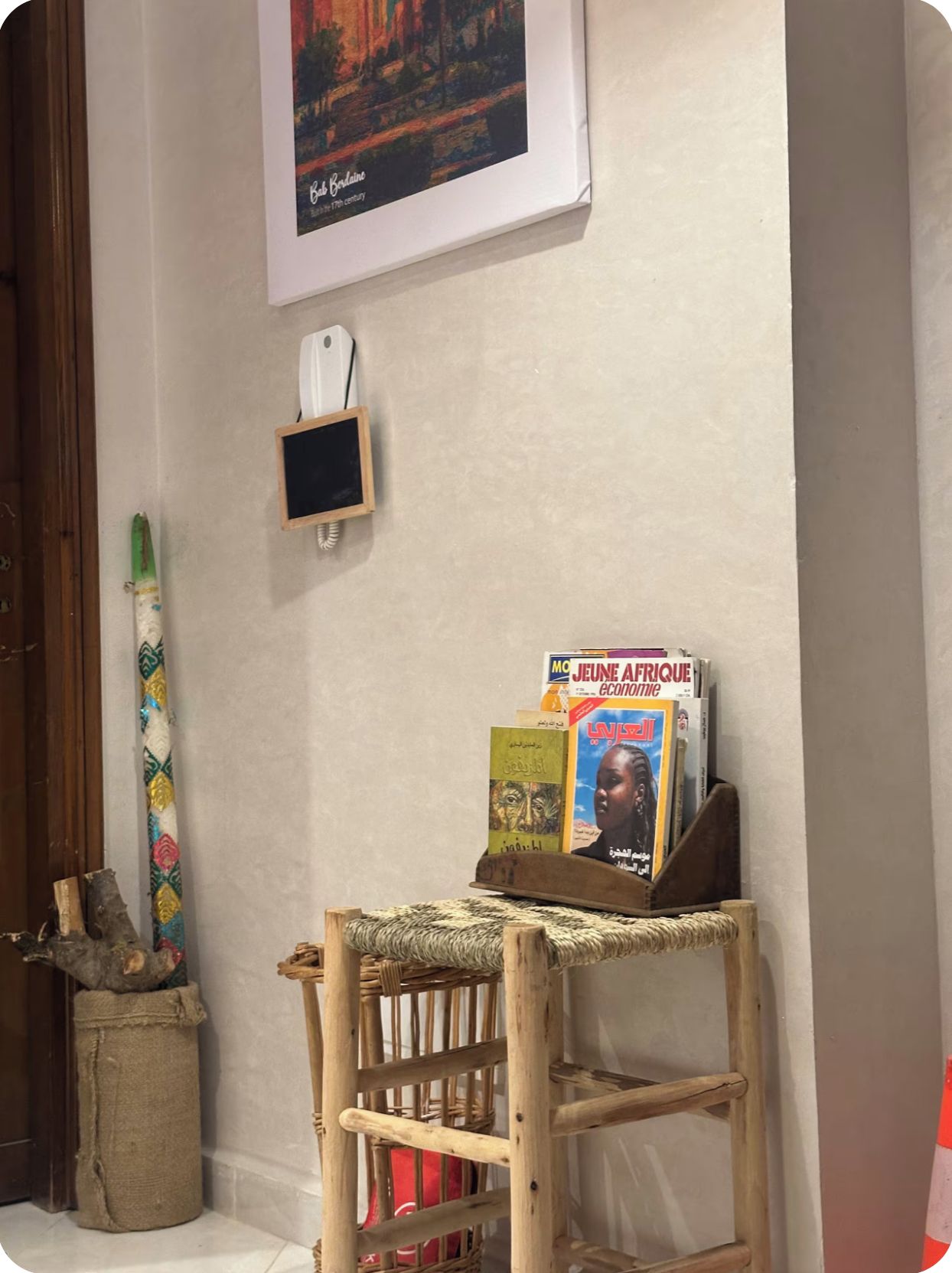 A corner of a room with a framed painting, a small blackboard, a wooden shelf with magazines, and a woven basket.