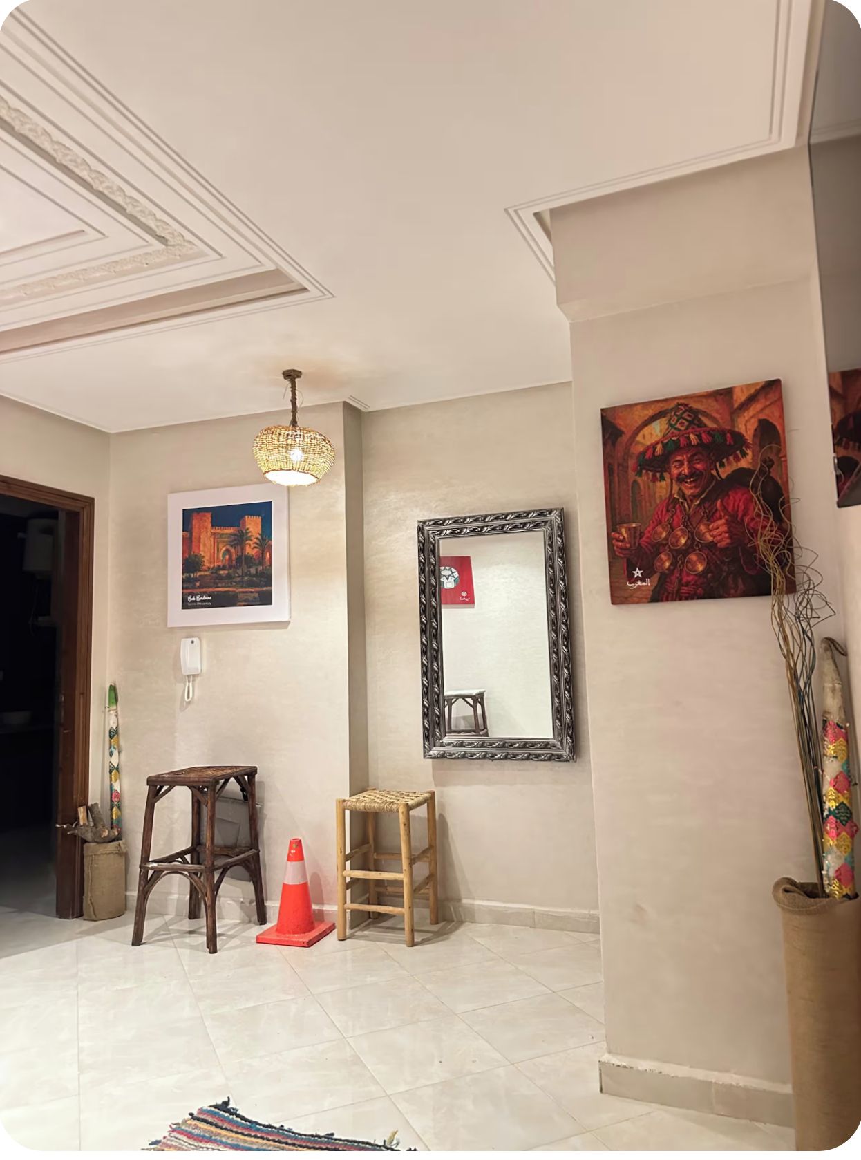 A room with a tiled floor, a wooden stool, a small table, a hanging mirror, and a painting on the wall.