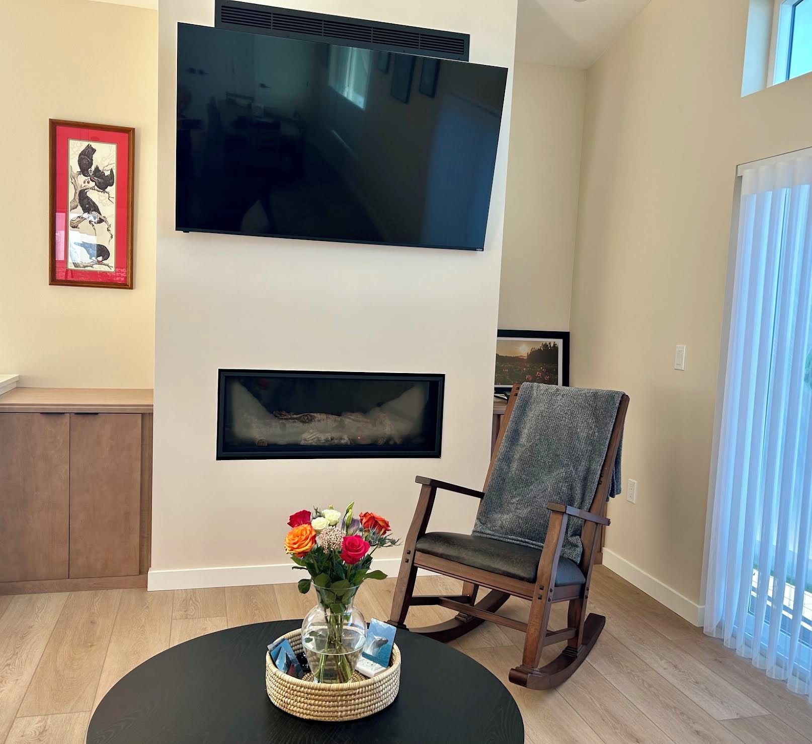 A living room with a flat-screen TV mounted on the wall, a fireplace below it, a wooden rocking chair, and a table with a vase of flowers.