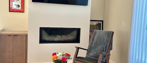 A living room with a flat-screen TV mounted on the wall, a fireplace below it, a wooden rocking chair, and a table with a vase of flowers.