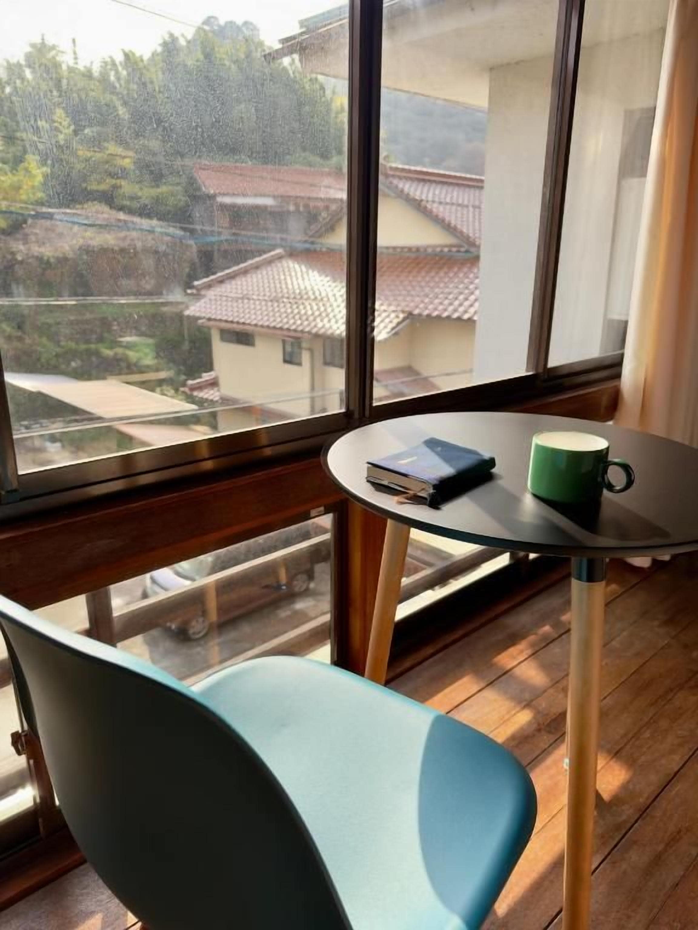 A room with a view of a residential area through a large window, a small round table with a book and a mug, and a blue chair.