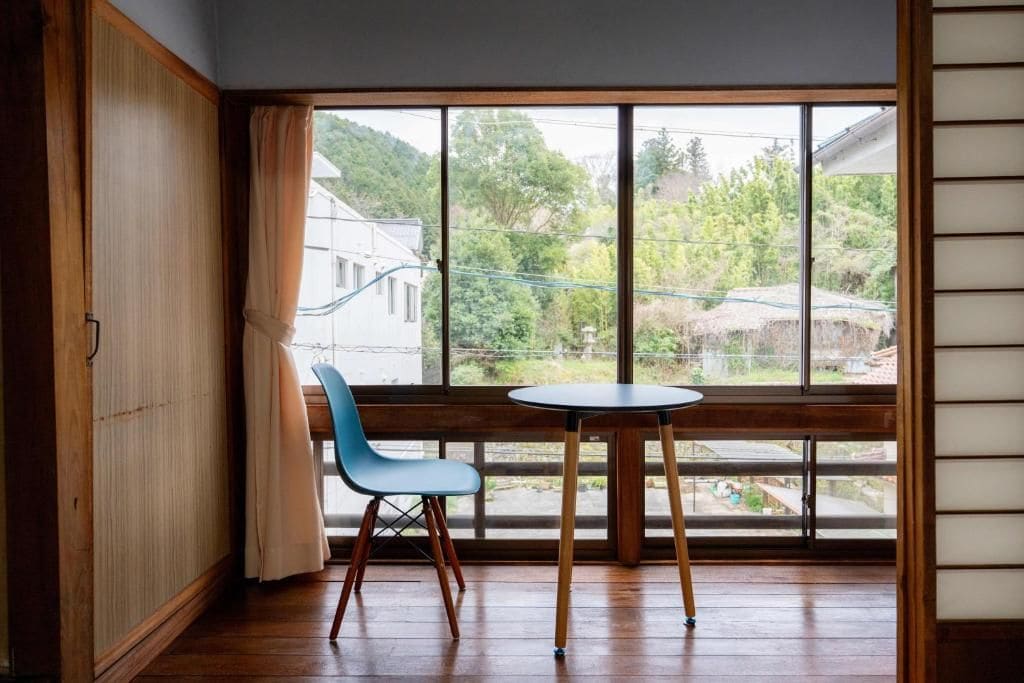 Une pièce avec un plancher en bois, une chaise bleue, une petite table ronde et une vue sur un quartier résidentiel, visible à travers de grandes fenêtres.