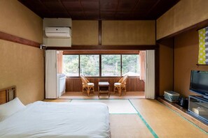 A room with a bed, a television, a window with curtains, and a view of greenery outside. - Tawarayama Machigoto Ryokan (Nagato)