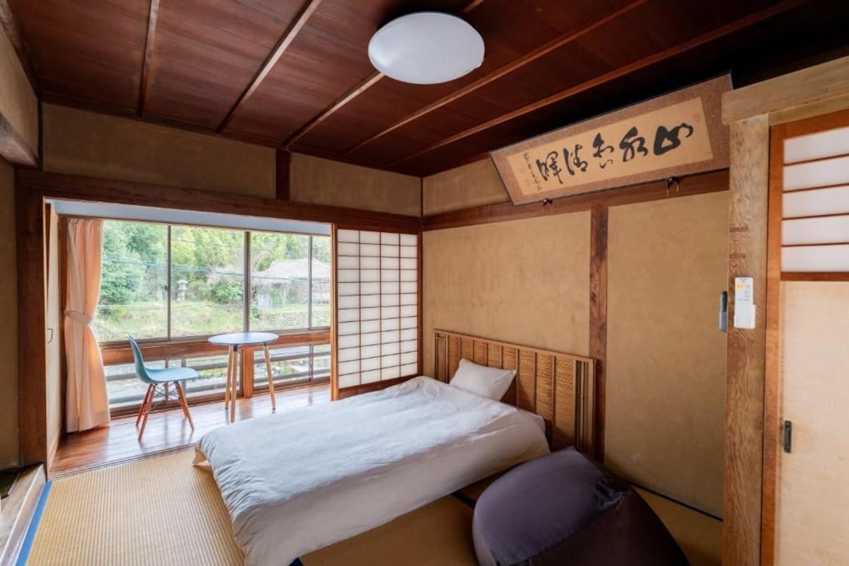 A traditional Japanese-style room with tatami flooring, a low wooden bed, a sliding shoji screen, and a view of greenery outside.