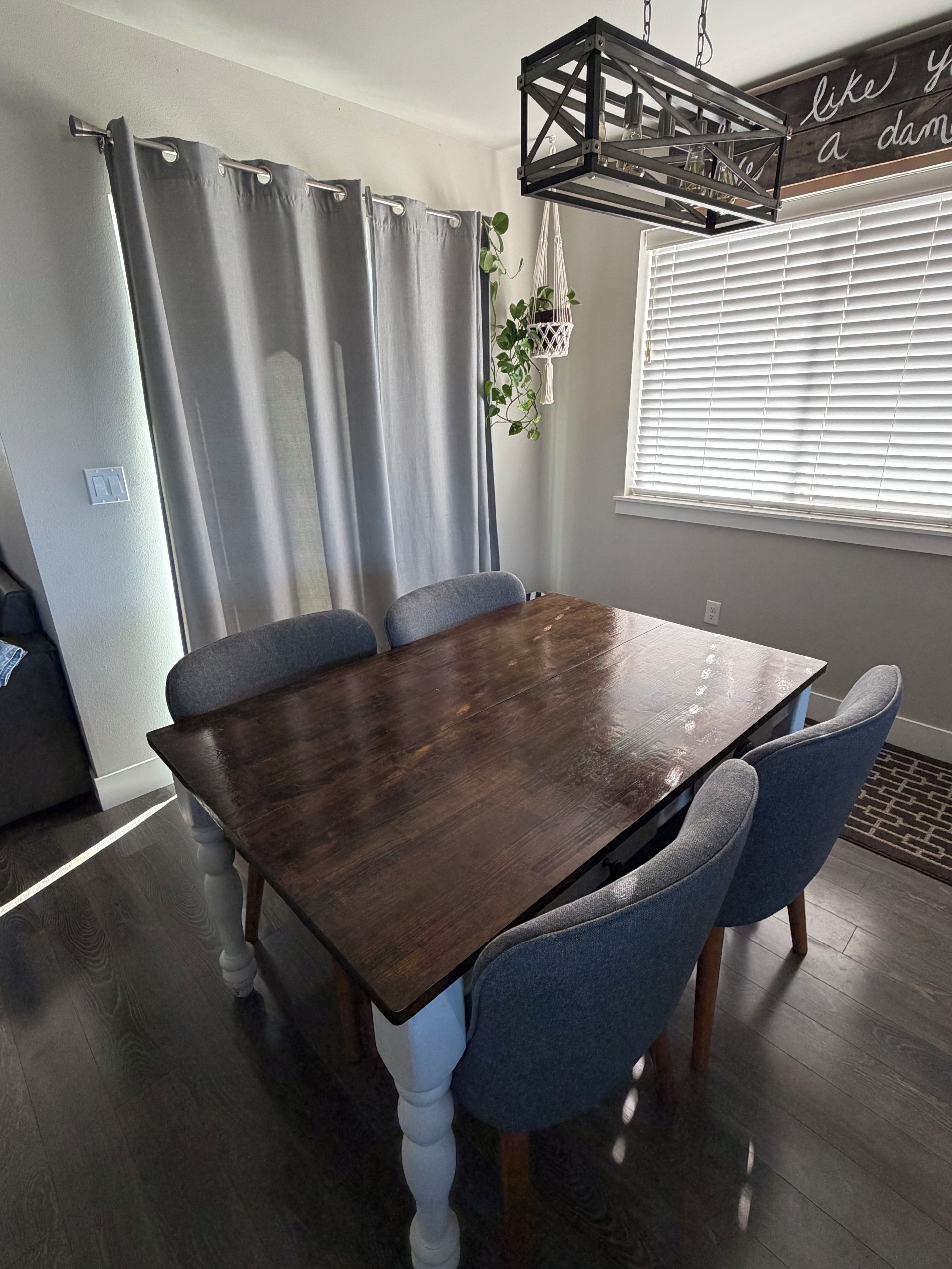 A dining area with a dark wooden table, four grey upholstered chairs, a grey curtain, and a hanging plant.