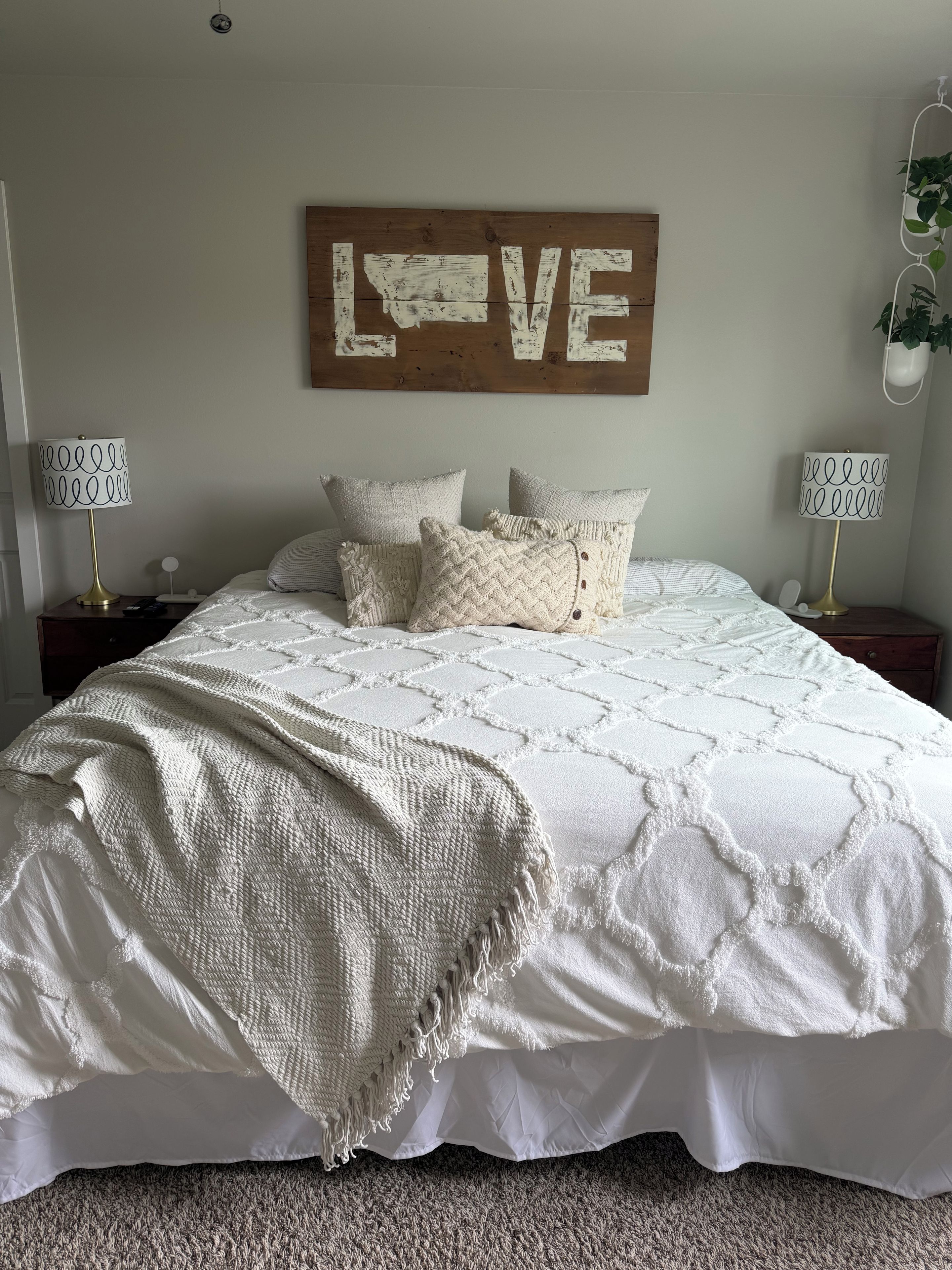 A bedroom with a large bed, two bedside tables, a wall-mounted sign, and a hanging plant.