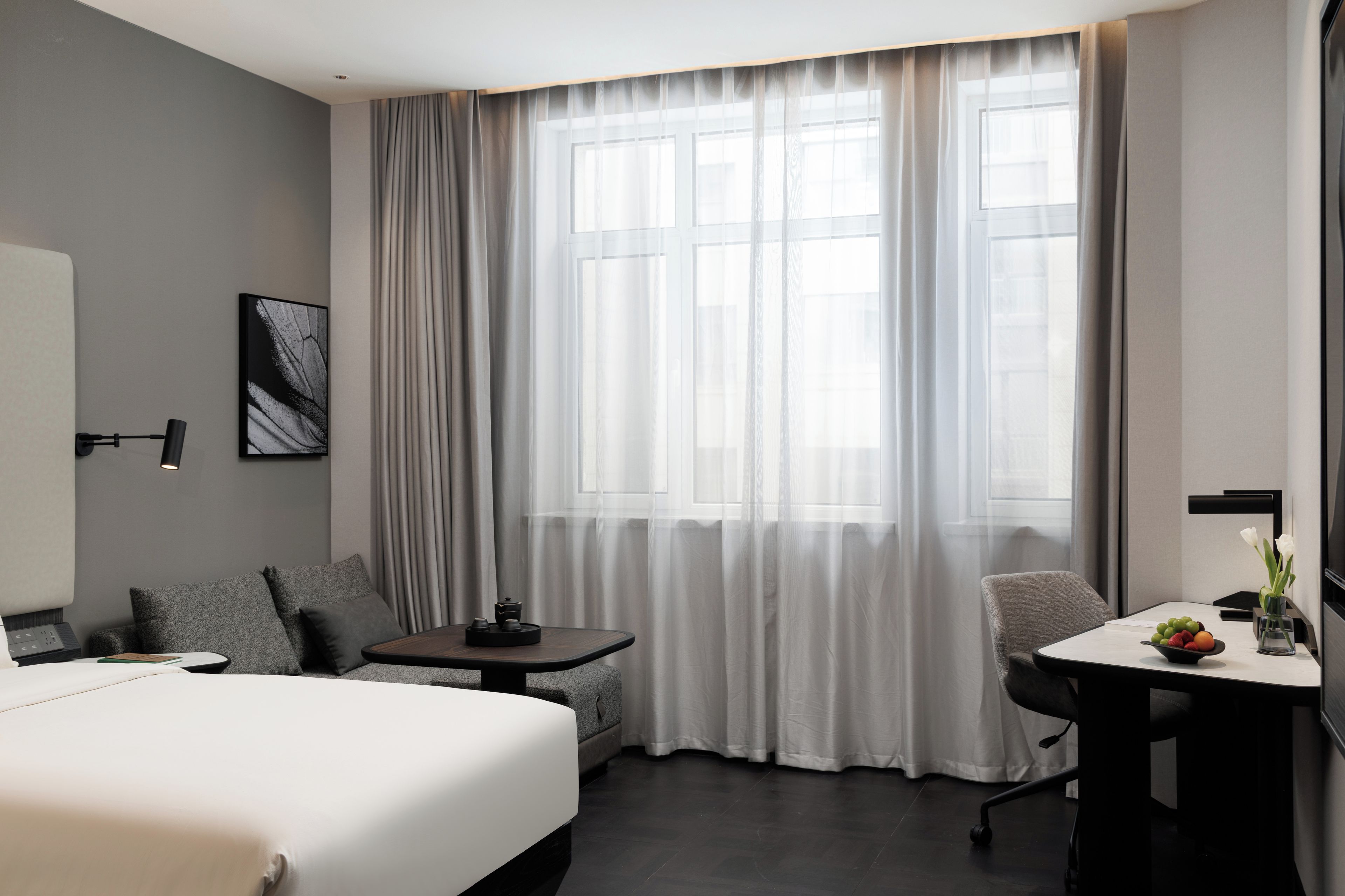 A hotel room with a bed, a desk, a chair, a small table with a fruit bowl, and a window with sheer curtains.