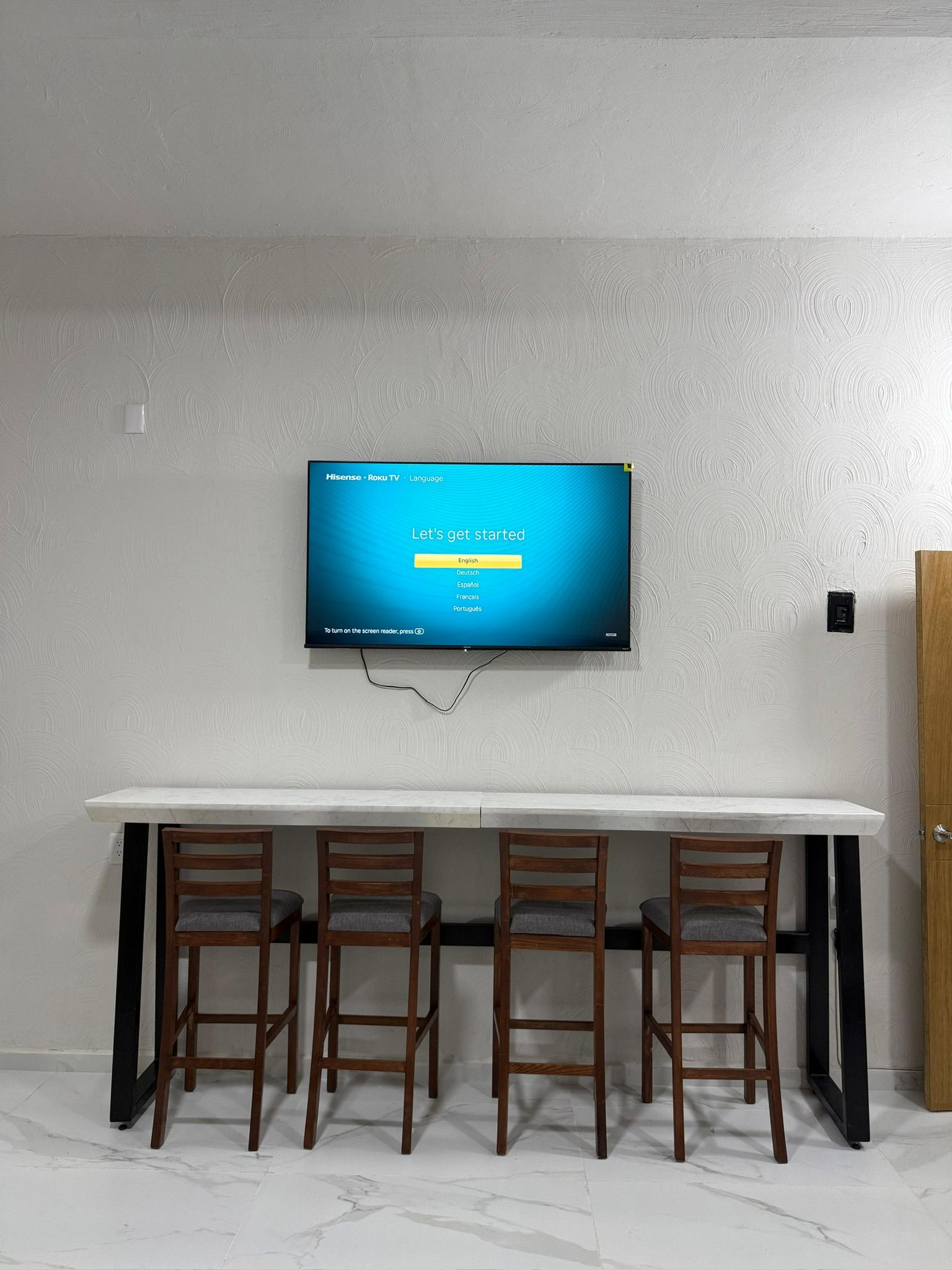 A flat-screen TV mounted on a wall above a white marble-topped table with four wooden chairs.