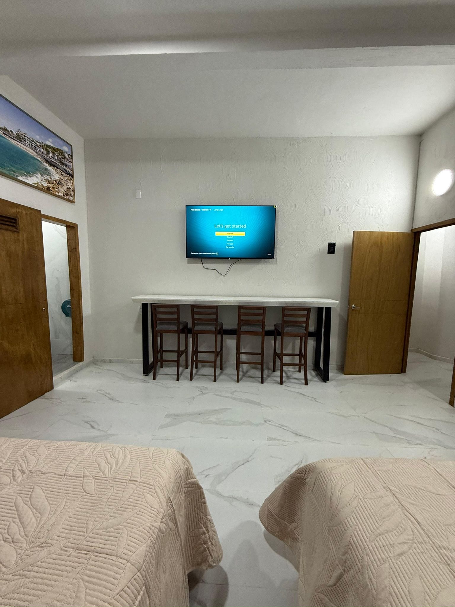 A room with a television mounted on the wall, a table with chairs, and two beds.