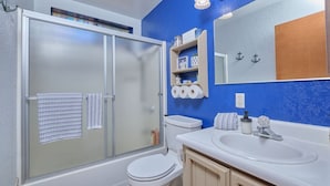 Un baño con cabina de ducha de vidrio, inodoro, lavabo con espejo y estante con artículos de aseo.