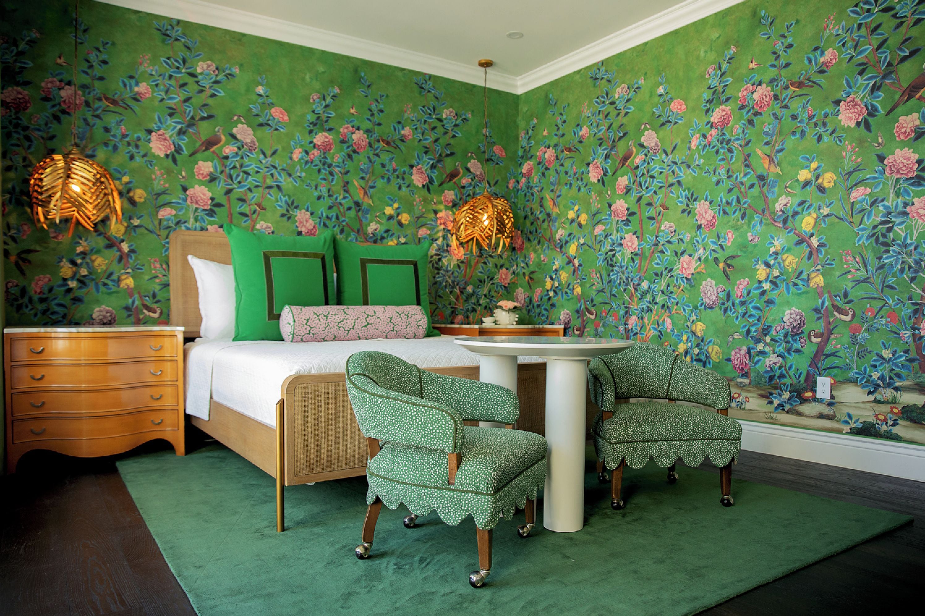 Un dormitorio con papel tapiz floral, una cama con ropa de cama verde y blanca, dos sillas, una mesita de noche y dos lámparas de pared.