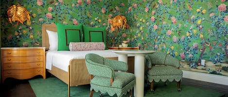 Un dormitorio con papel tapiz floral, una cama con ropa de cama verde y blanca, dos sillas, una mesita de noche y dos lámparas de pared.