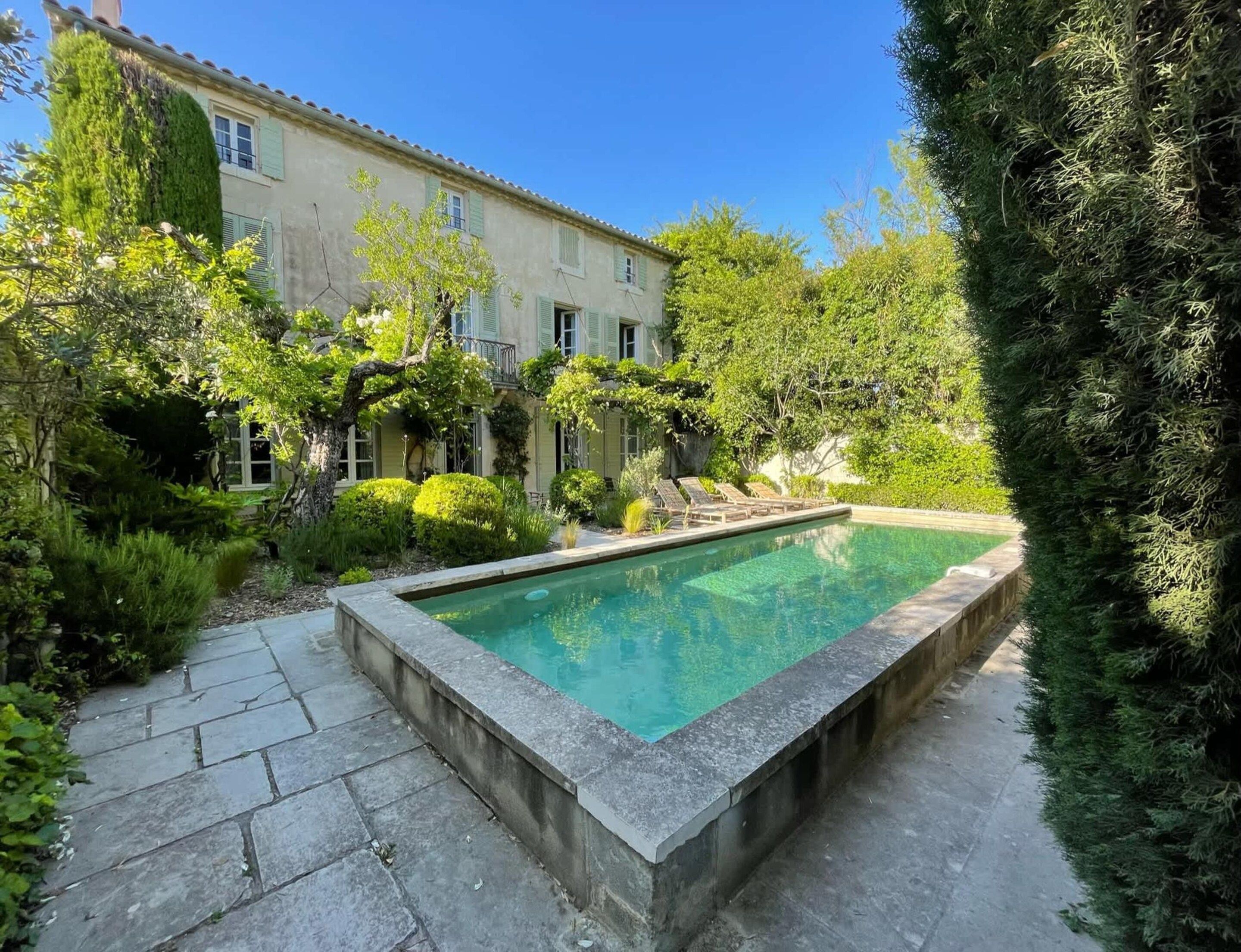 A rectangular swimming pool with a stone border, surrounded by a stone pathway, and a building with a tiled roof in the background.