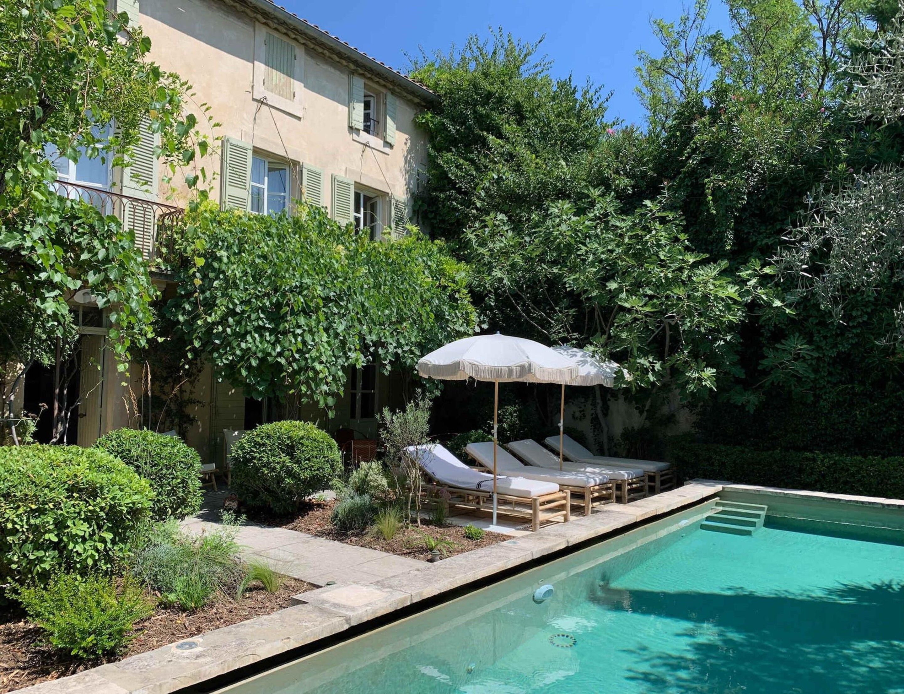 A poolside area with lounge chairs and an umbrella, surrounded by greenery and a building.