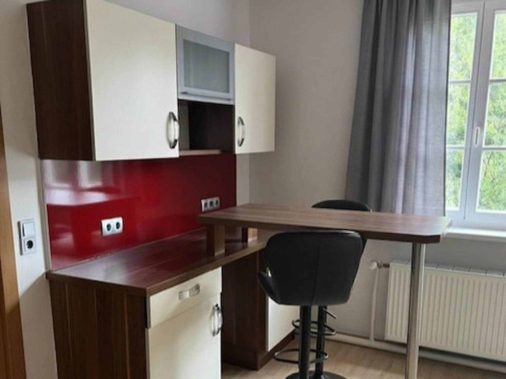 Une petite cuisine équipée avec un plan de travail rouge, des placards blancs et une petite fenêtre agrémentée de rideaux.