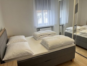 Ein Einzelbett mit grauem Kopfteil und weißer Bettwäsche in einem Zimmer mit Fenster, Heizkörper und Spiegel.