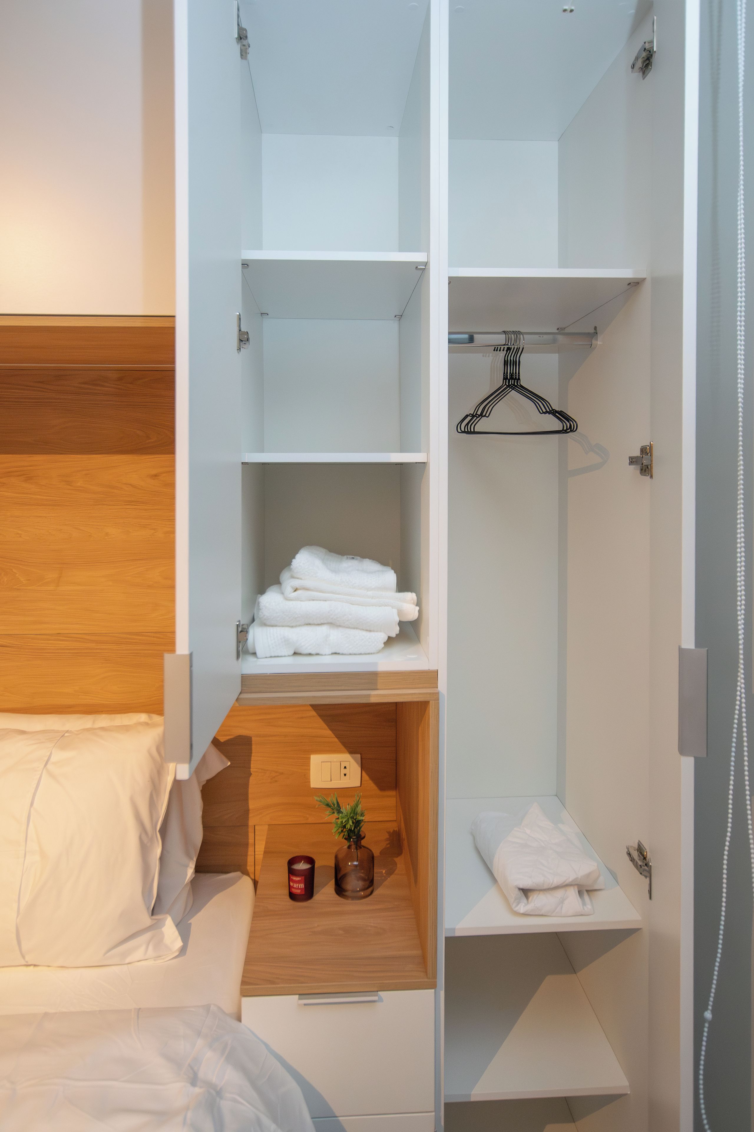 Ein Hotelzimmer mit einem hölzernen Kopfteil, einem Bett mit weißen Leinentüchern, einem Nachttisch mit Kerze und kleiner Pflanze sowie einem weißen Kleiderschrank mit Regalen und Kleiderhaken.