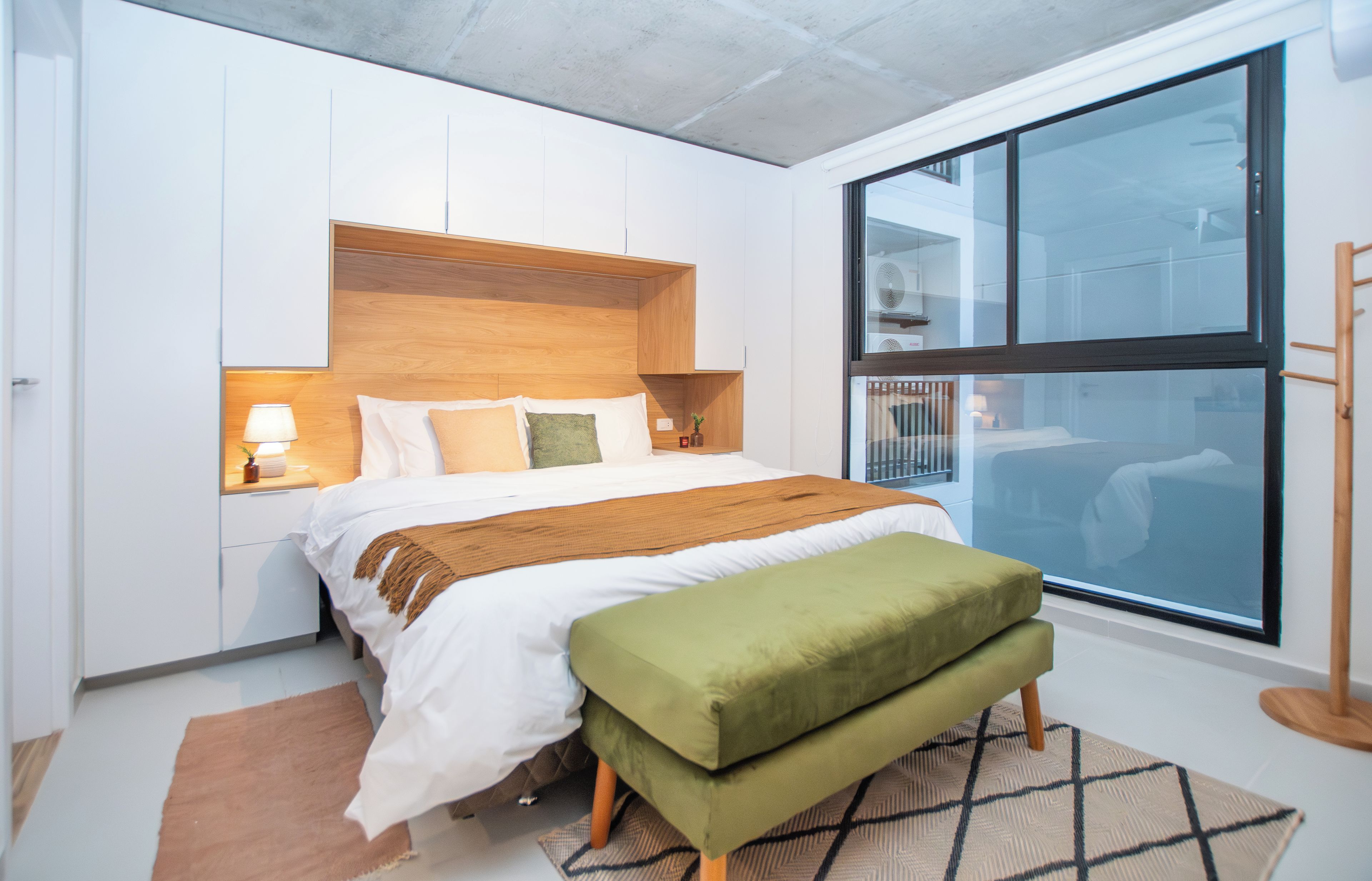 A modern bedroom with a large bed, wooden headboard, bedside lamps, a bench, and a large window.