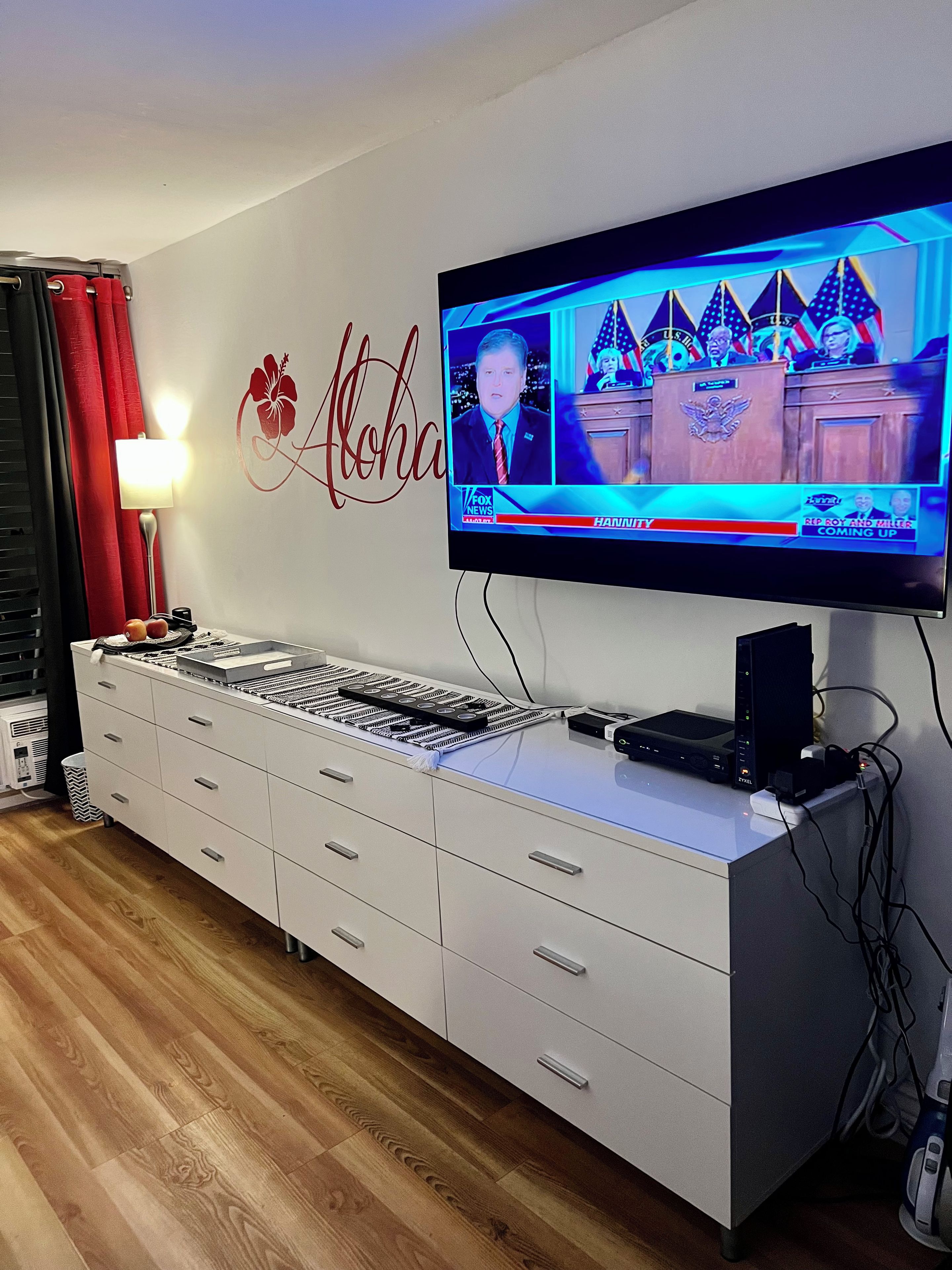 Ein Zimmer mit Fernseher, der einen Nachrichtensender zeigt, einem weißen Schrank mit Schubladen, einer Lampe und einer Wand mit Blumenmuster.